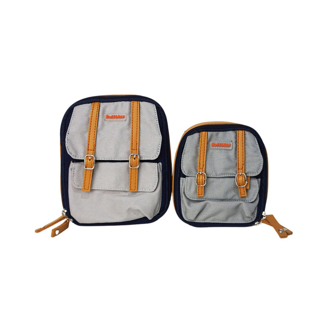 Dale Navy model Ruffsack backpack front angle. The backpack face features a cotton gray solid pattern with two small front buttoned pockets atop a cotton navy blue solid pattern. Tan faux leather accents are on the front and sides of the bag. A side zipper allows access to the inside.